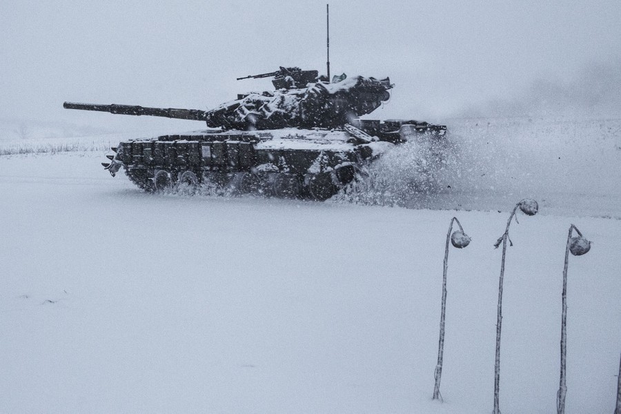 A tank moves along a snow-covered road, kicking up clumps of snow.