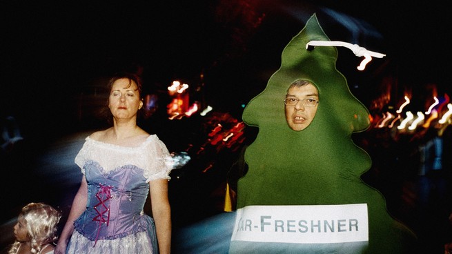 A man dressed in an air-freshener tree costume and a woman dressed as a princess walk amidst blurred bright lights