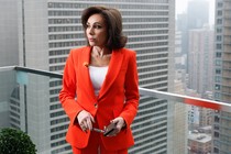 Jeanine Pirro wearing an orange suit and standing on a balcony with a metal and glass railing in front of a cityscape