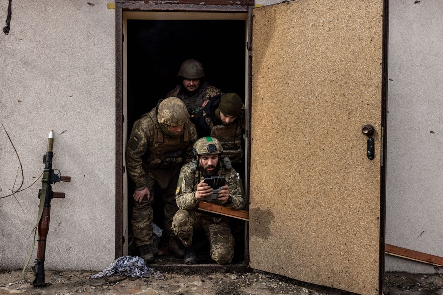 Four soldiers gather in a doorway, looking on while one operates a remote control for a drone.