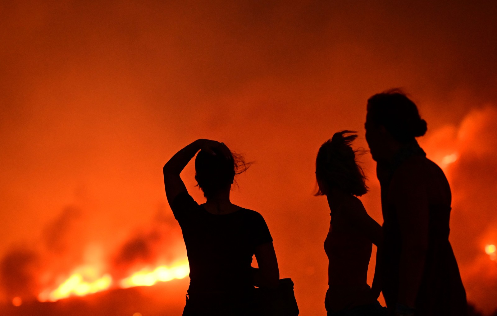 Several people are seen in silhouette, looking toward a wildfire at night.