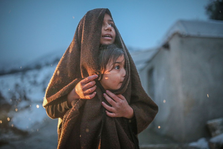 Two young children share a blanket while standing in a refugee camp in cold weather.