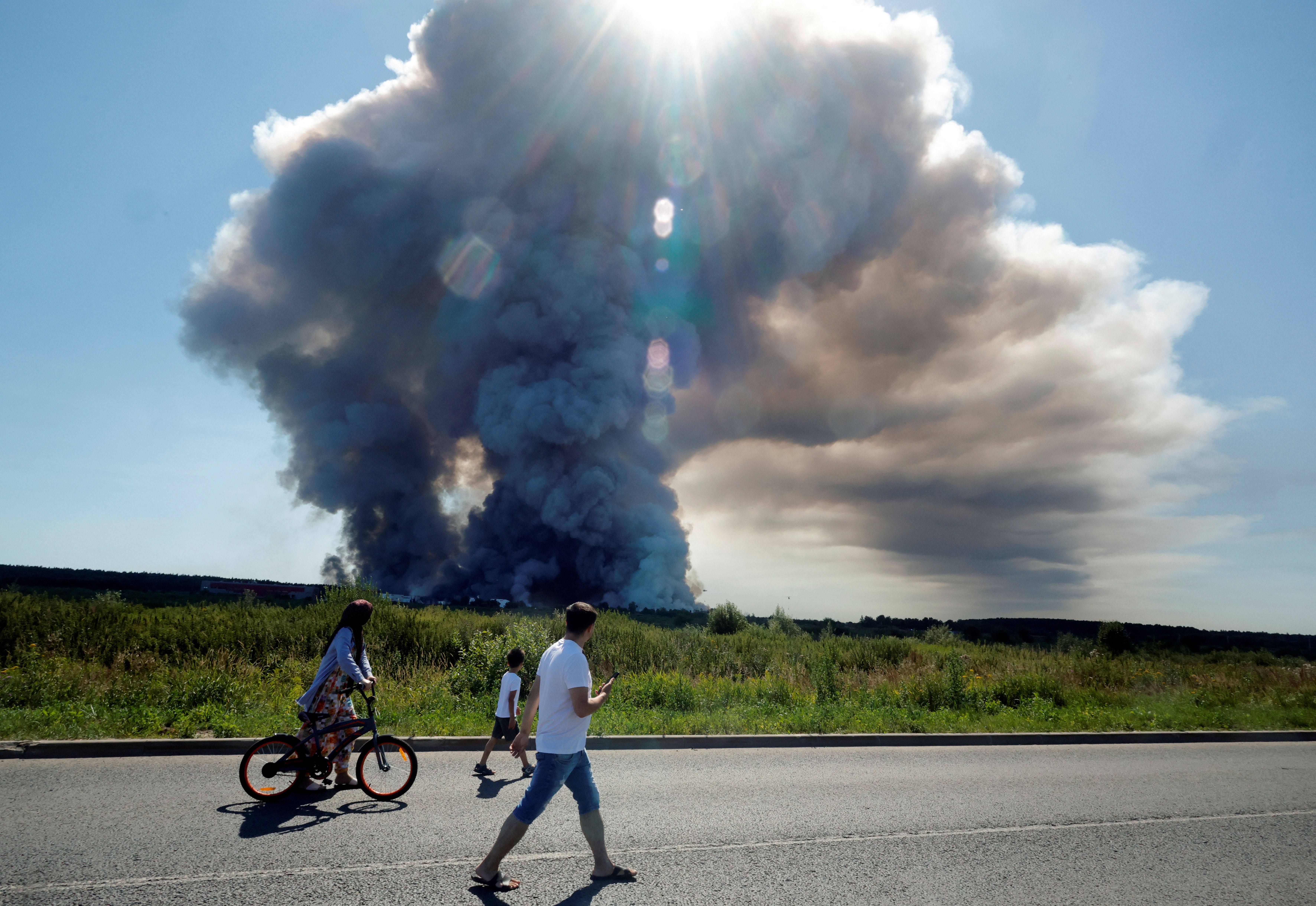 Several people walk on a road past the smoke of a fire burning in the distance.