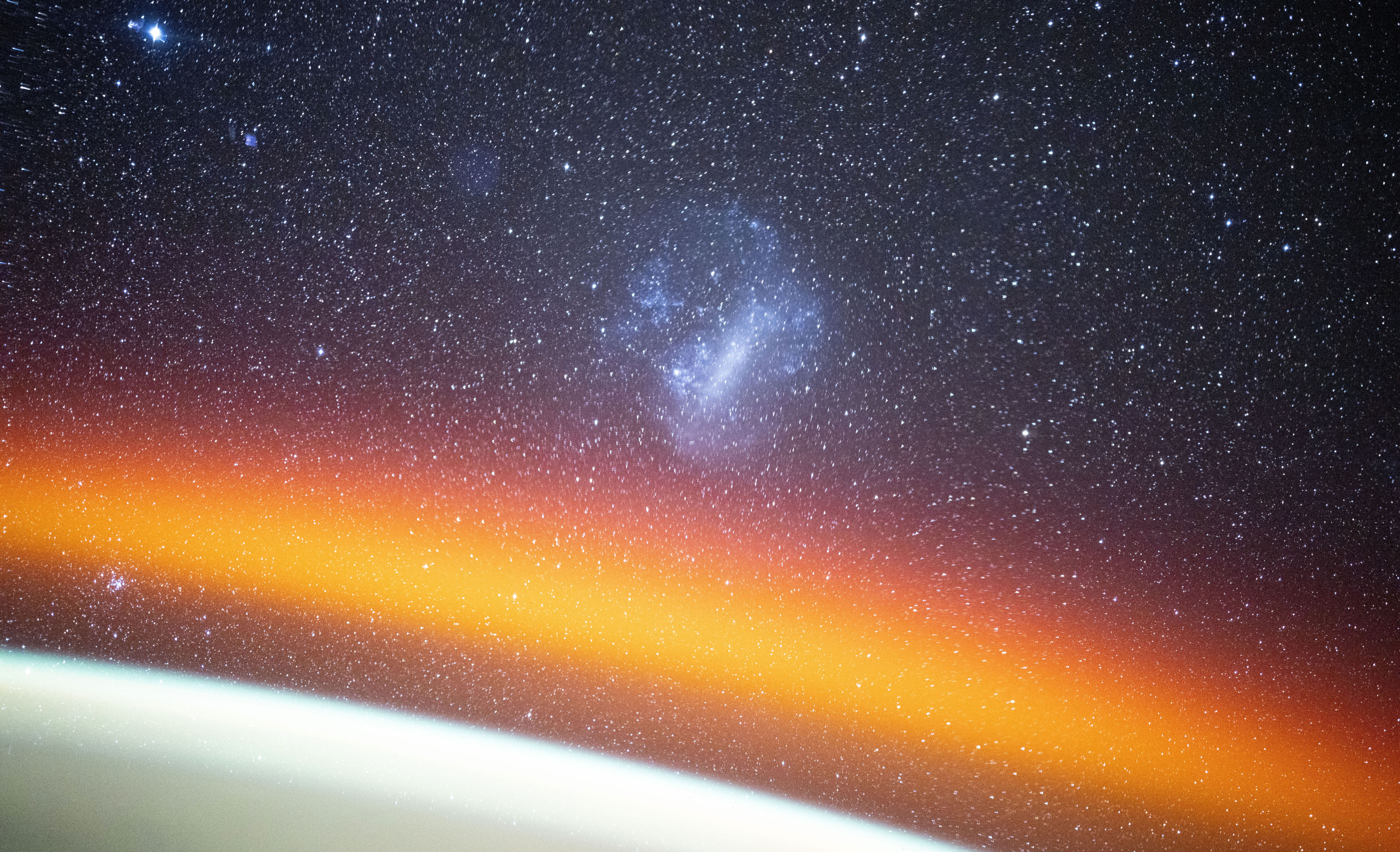 An orange glow appears above the Earth's horizon, seen from orbit, with stars and a small irregular galaxy in the background.
