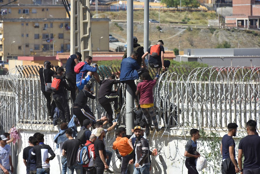 About a dozen people climb over a fence topped by razor wire.