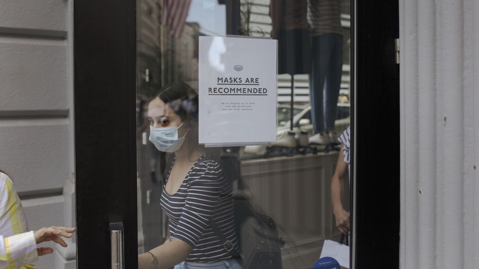 A woman wearing a mask opens a door. On the door is a sign that reads "Masks are recommended."