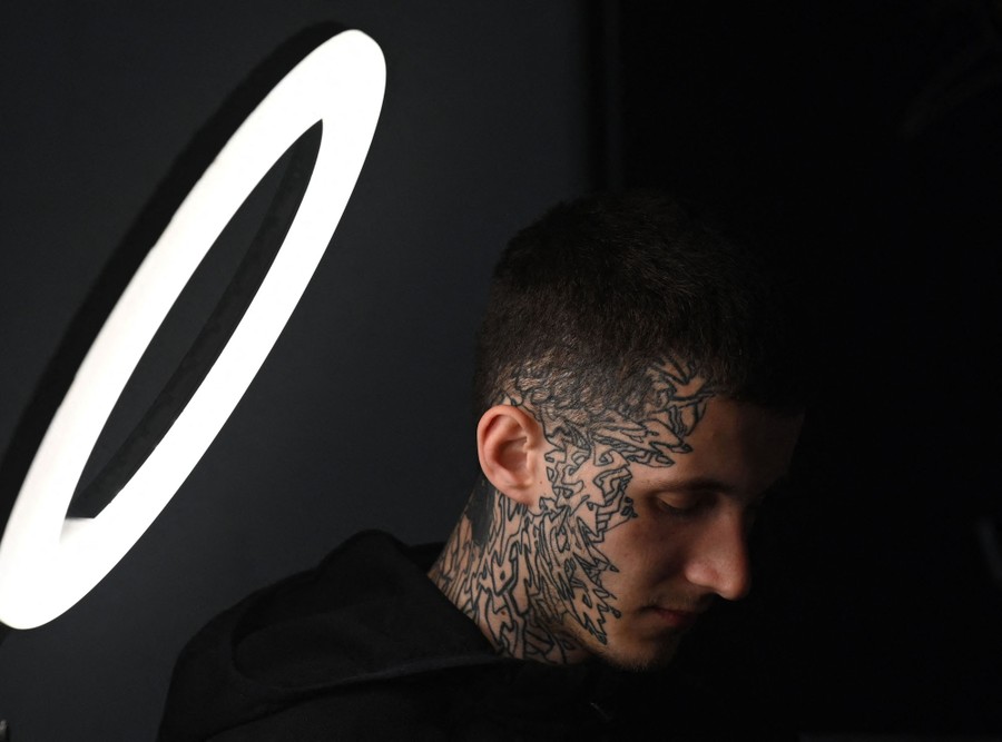 A man sits beside a lamp, with an intricate tattoo on his face and scalp.