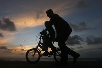 A man guides a child riding a two-wheel bike. It's sunset, and the figures appear as silhouettes.