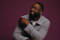 photo of Dwyane Wade smiling and sitting with arms crossed, wearing gray collared jacket, against purple background