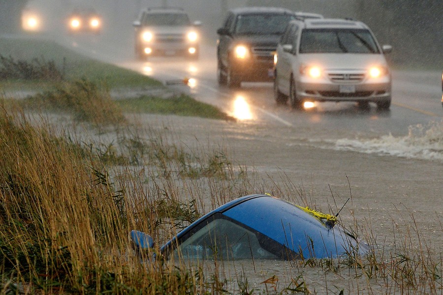 Cars on a partially-flooded road pass by a car in a ditch that is submerged to its roof.