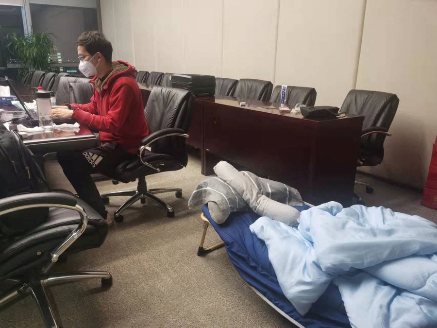 A person works at a laptop in an office with a small portable bed on the floor beside them.