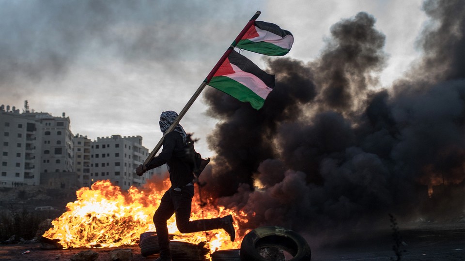 A man holding a Palestinian flag runs in front of a blazing fire.