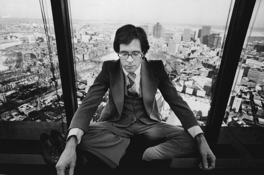 A man wearing a business suit sits cross-legged in the window of a tall building, with Boston visible behind him.