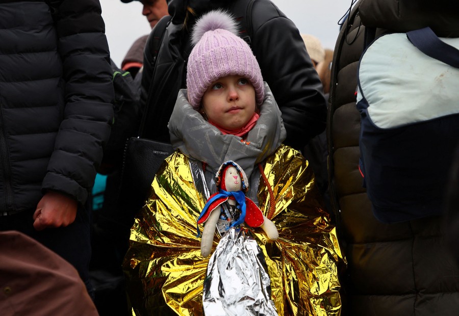 A young child wearing a shiny, reflective rescue blanket holds a doll while standing with a crowd of adults.