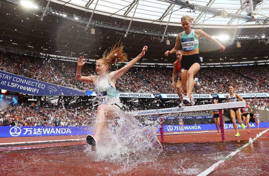 Runners on a track jump over a barrier into a shallow pool of water during a race.