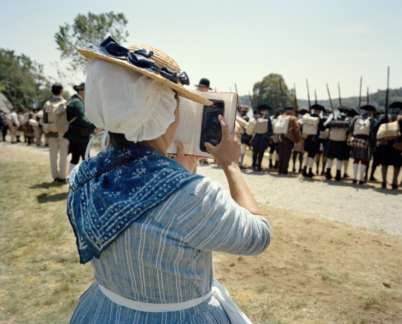 photo of female reenactor dressed in period blue dress, kerchief, and bonnet holding up a book that disguises the cell phone she's looking at behind it, with a row of soldiers with muskets facing away in the background