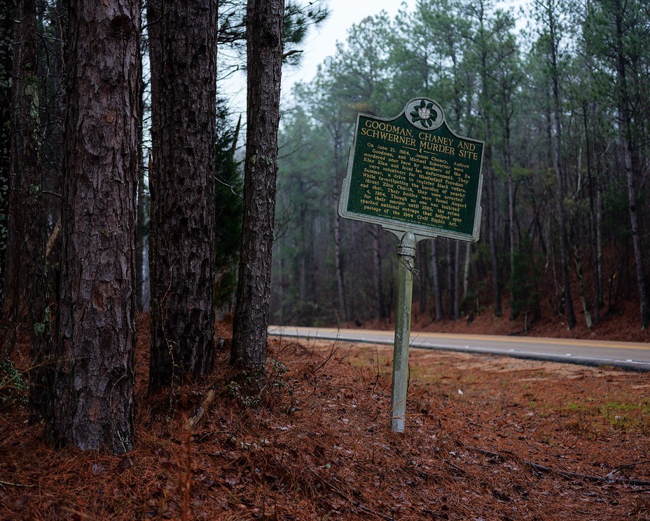 photo of roadside memorial marker including text Goodman Chaney and Schwerner Murder Site next to trees with road and forest in background