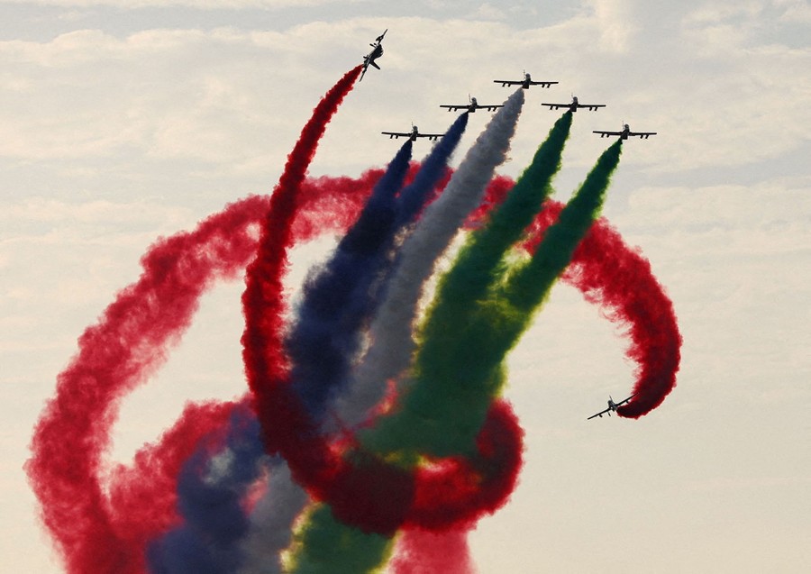 Five small aircraft trailing colored smoke fly side-by-side, as two others spiral around them all.