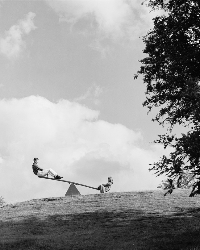 a black and white photograph of a father and daughter on a see saw next to a large tree