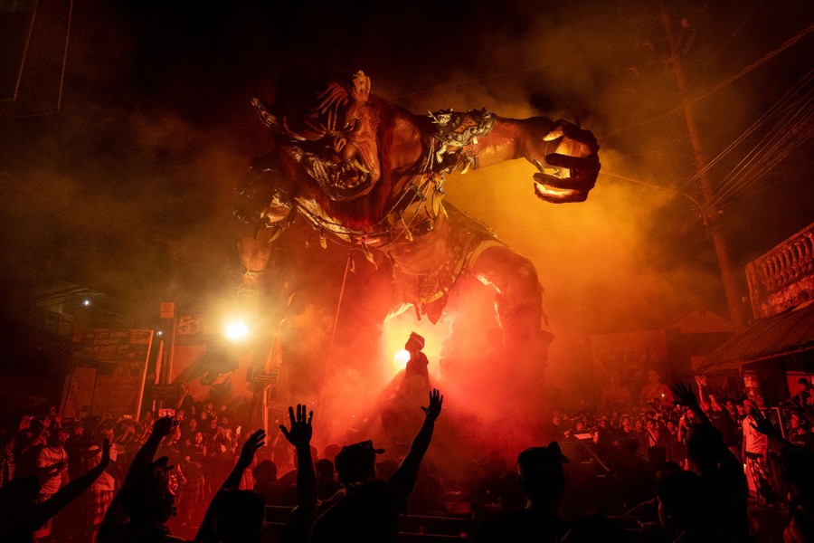 A crowd gathers in a street at night, around a giant sculpture of a demon, during a festival.
