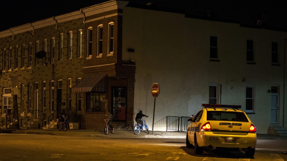 A police car sits on a street in East Baltimore, shining its headlights on a man riding a bike.