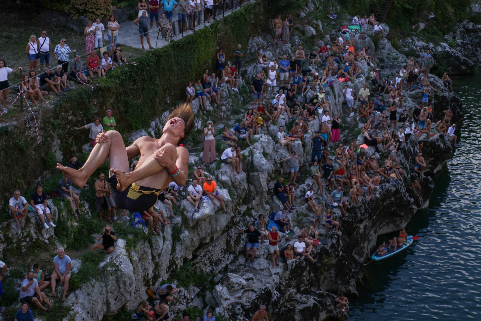A diver spins in mid-air, as they drop toward a river below, watched by a crowd.