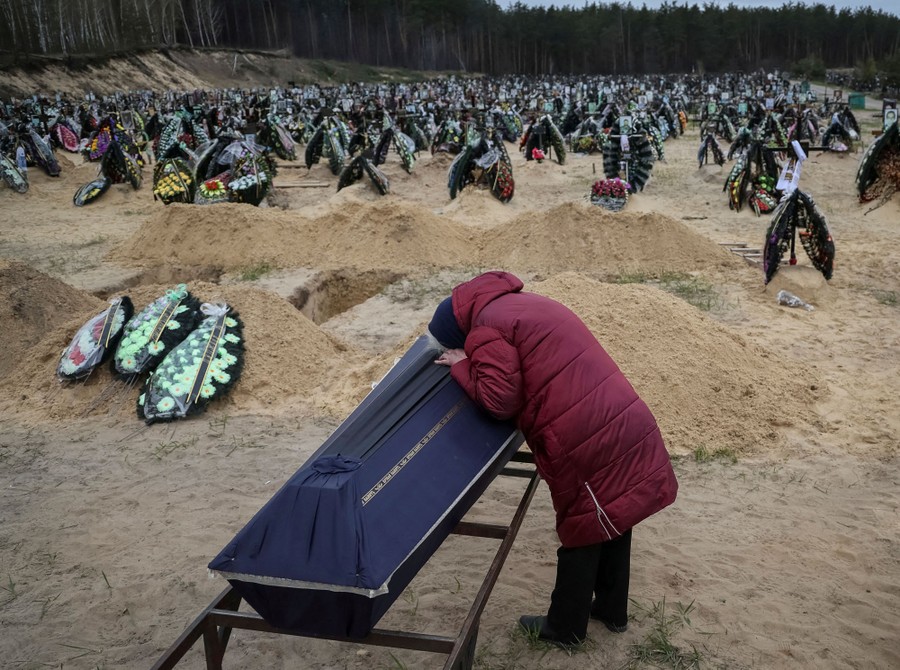 A person leans over a coffin in a cemetery near newly dug graves.