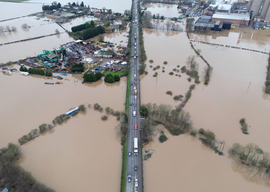 Cars and trucks drive on a road surrounded by flooded fields.