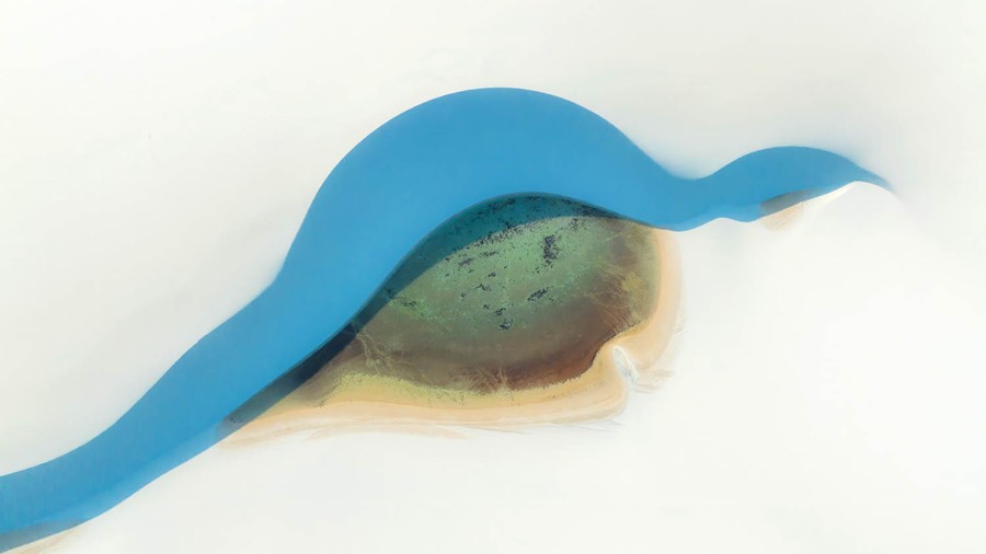 An aerial view of a thin strip of blue water in a small valley among sand dunes, with a rounded, darker-colored bank at bottom that makes the whole scene look like a stylized painting of an eye.