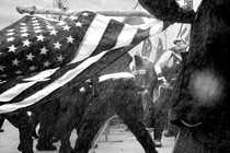 A black-and-white photo of an American flag held sideways in front of a row of cops facing off against men in tactical vests and MAGA sweatshirts.