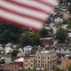 Color photograph of the town of Charleroi seen at a distance on what appears to be an overcast day. A blurry American flag whips across the top of the frame at close range.