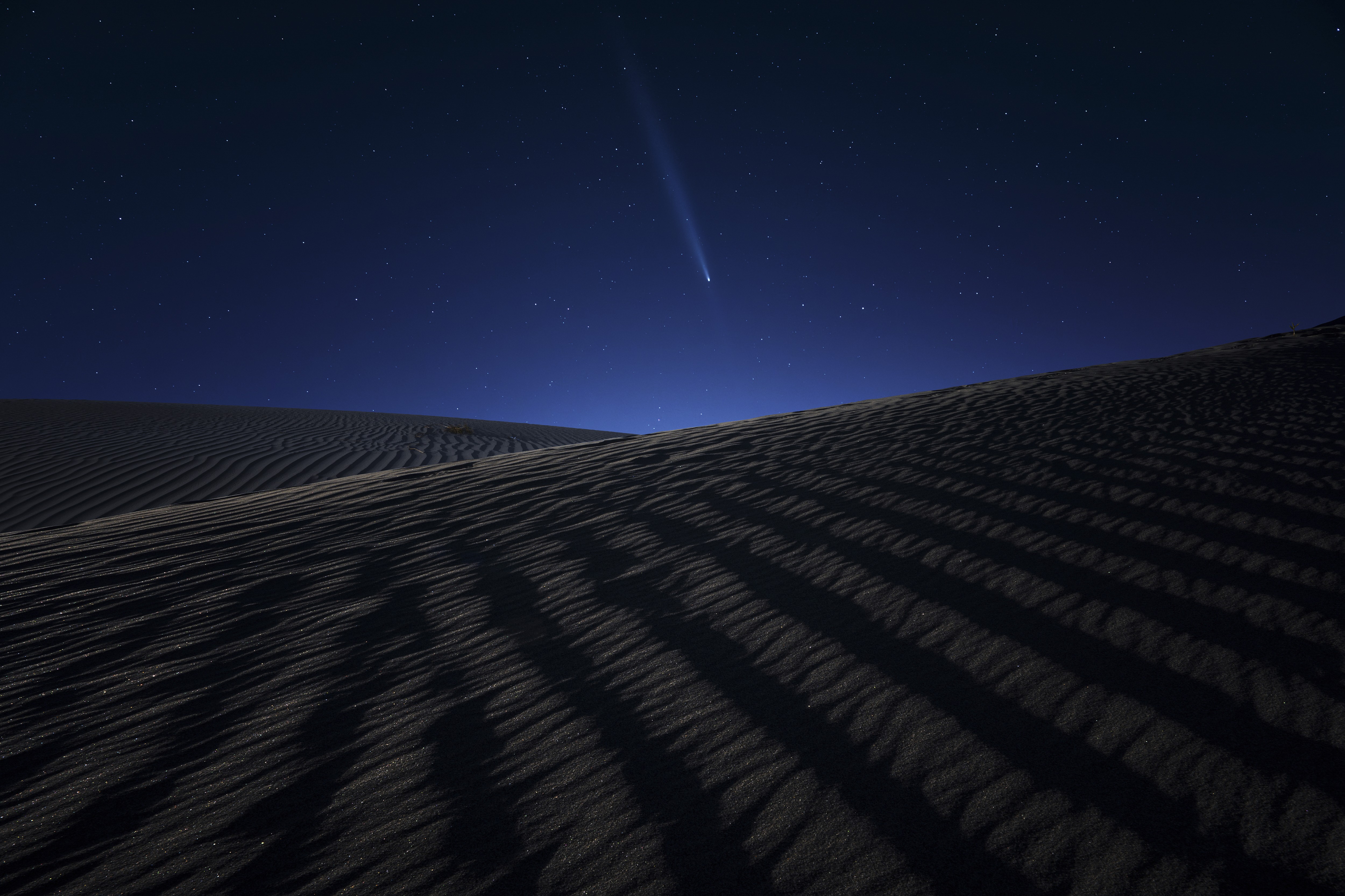 A night view of a comet in the sky above broad rippled sand dunes