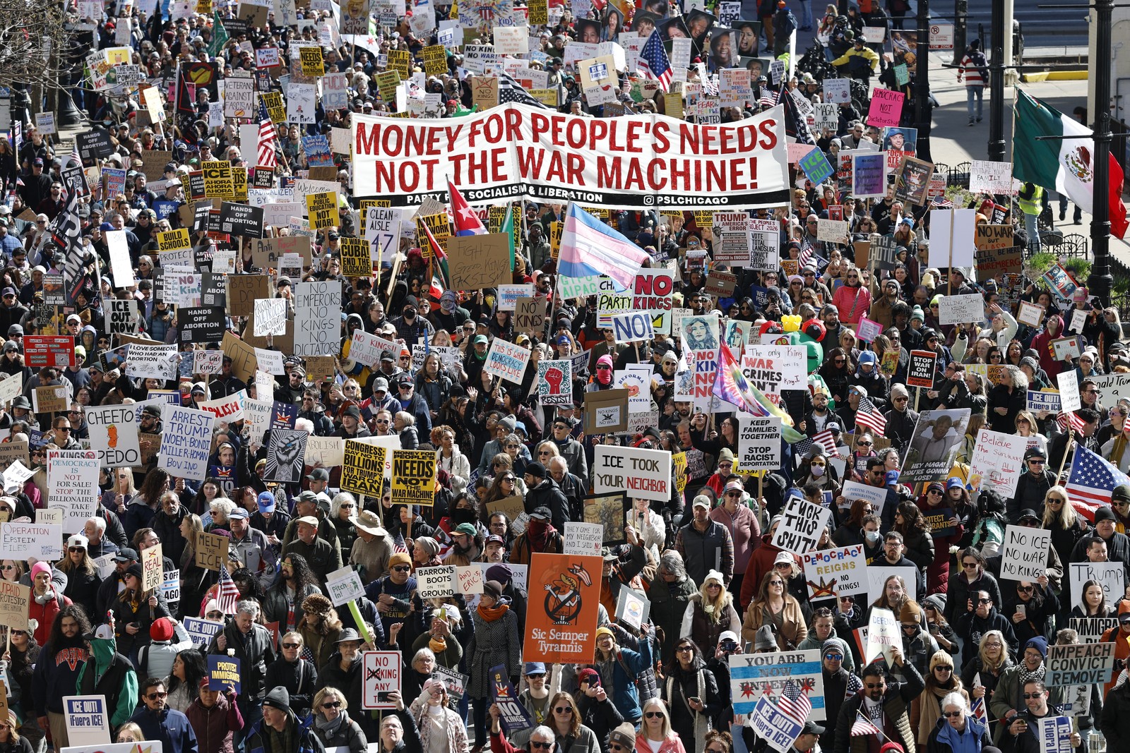 A large crowd of protesters marches down a city street carry many signs and a banner that reads 