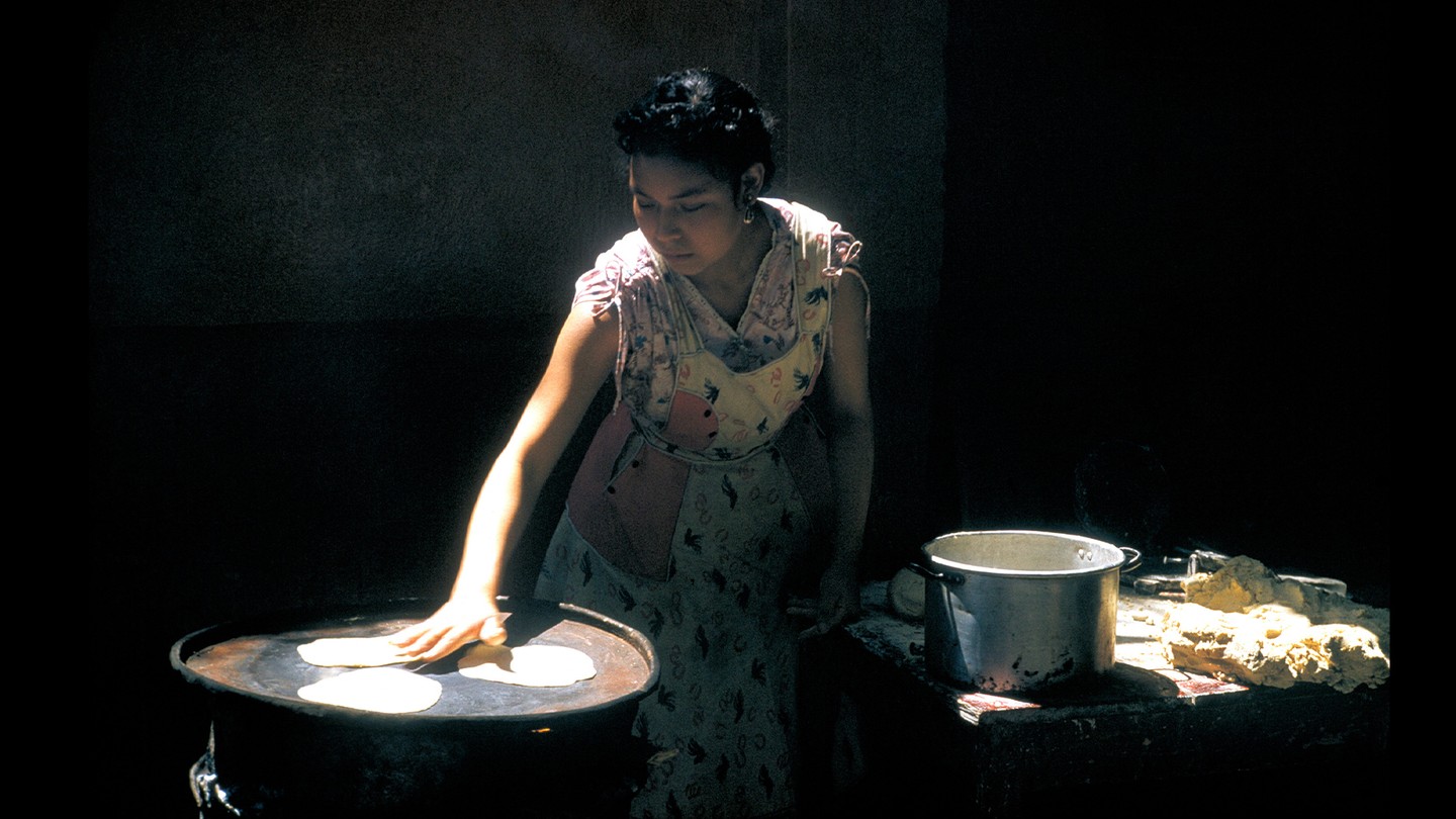 A woman lit by a ray of light against a black background lays out tortillas she's cut from dough