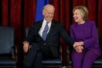 Joe Biden and Hillary Clinton sit on chairs laughing together in front of a red curtain.