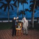 Two men standing in front of a row of palm trees at night giving a speech, alongside an empty chair.