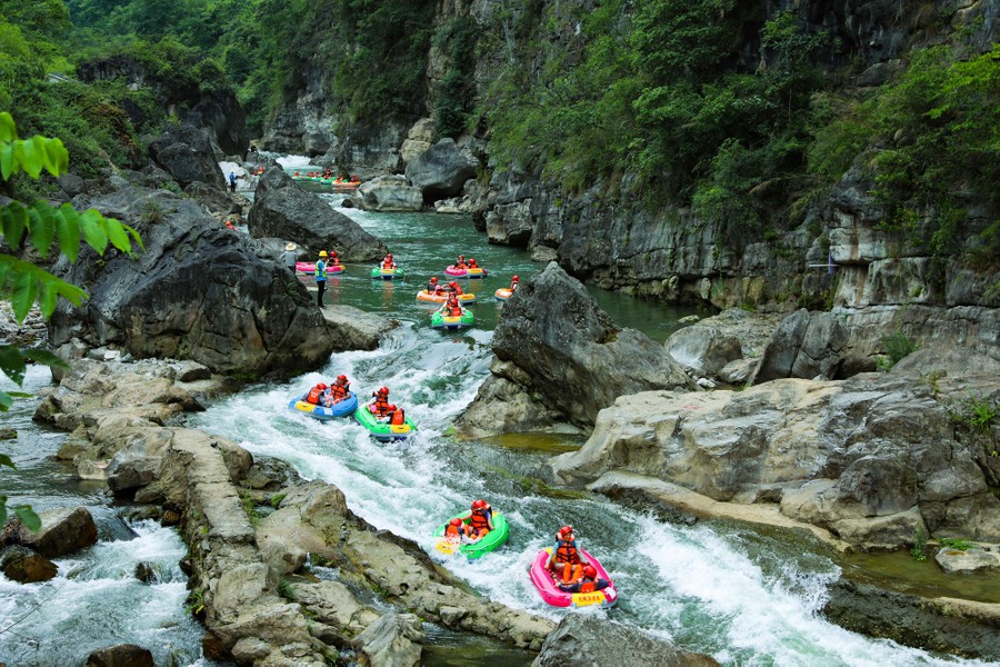 More than a dozen colorful rafts carry tourists down a mild set of rapids in a steep river valley.