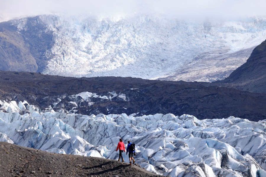 Two visitors walk on a ridge in front of a large glacier.