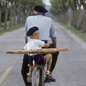 An old man and a young boy, both in berets, ride a bicycle with two baguettes strapped to the back down a tree-lined street; the young boy is looking back over his shoulder at the camera.
