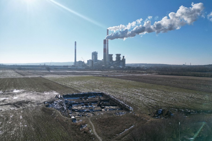 An elevated view of the excavated site of an ancient Roman structure in an open field with a power plant in the background