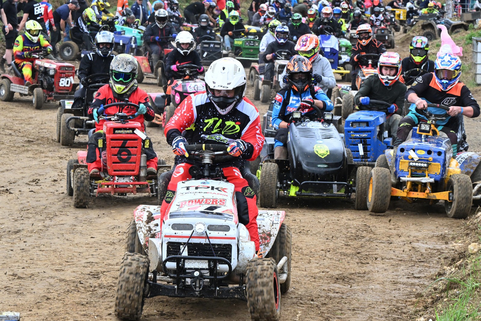 Dozens of helmeted competitors race, driving lawn tractors on a dirt track.