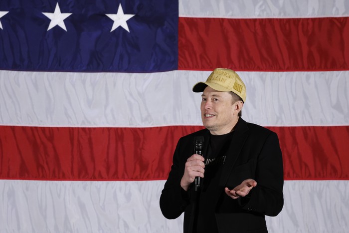 Elon Musk shrugs wearing a MAGA hat in front of an American flag