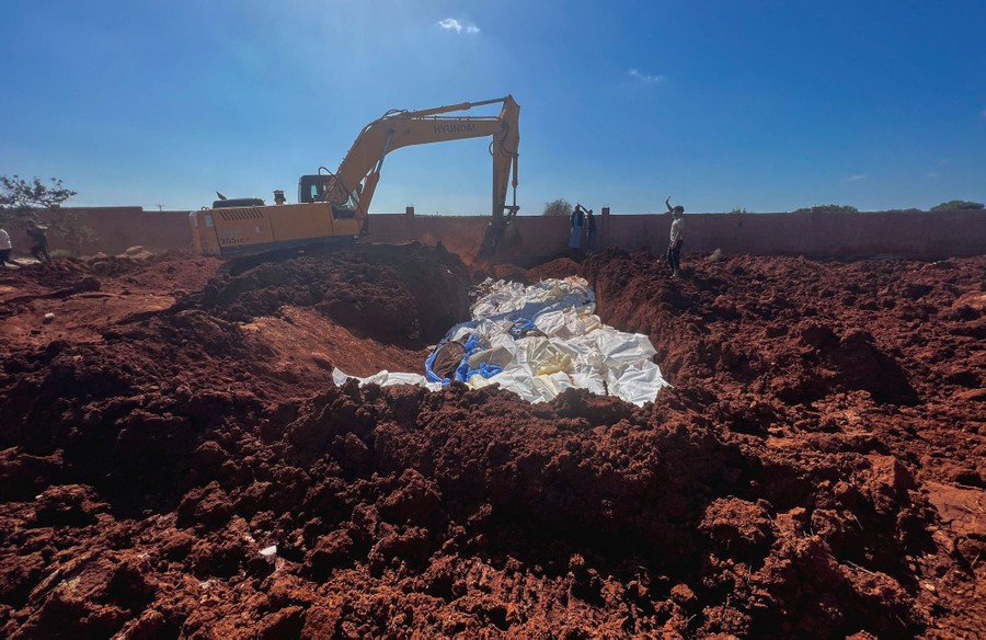 An excavator sits beside a broad pit that has been filled with dozens of bodies wrapped in fabric.
