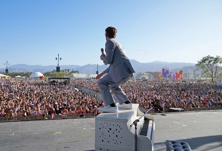 A singer stands on top of a piano onstage during an outdoor concert.