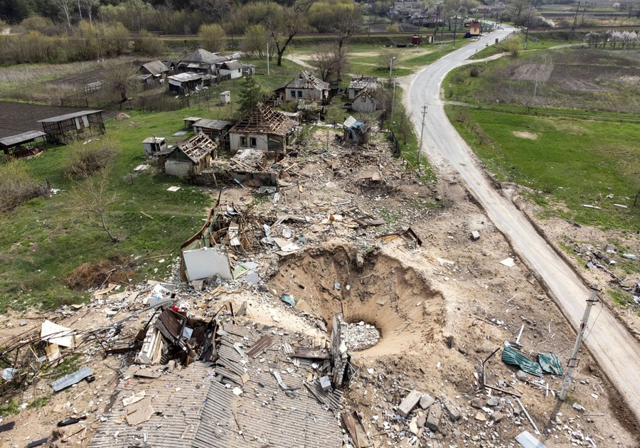 An aerial view of a crater and the debris of several destroyed buildings, in a rural area.