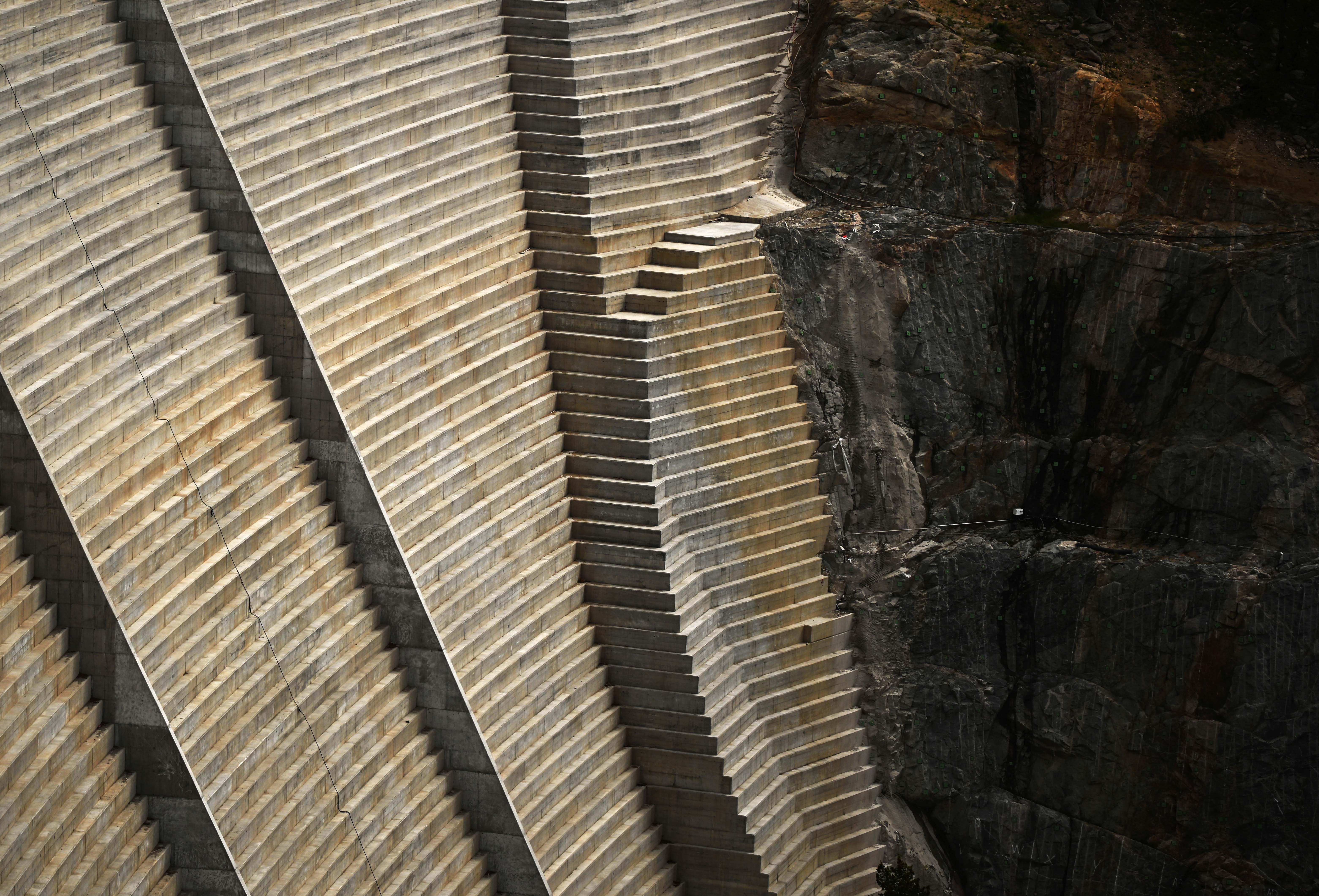 A steep, stepped wall of concrete—part of a huge dam—seen against a stone hillside