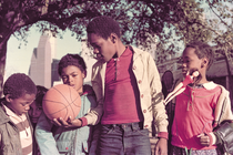 color photo of 5 children, one holding out basketball and another a Barbie doll, under large tree with Houston skyline in background