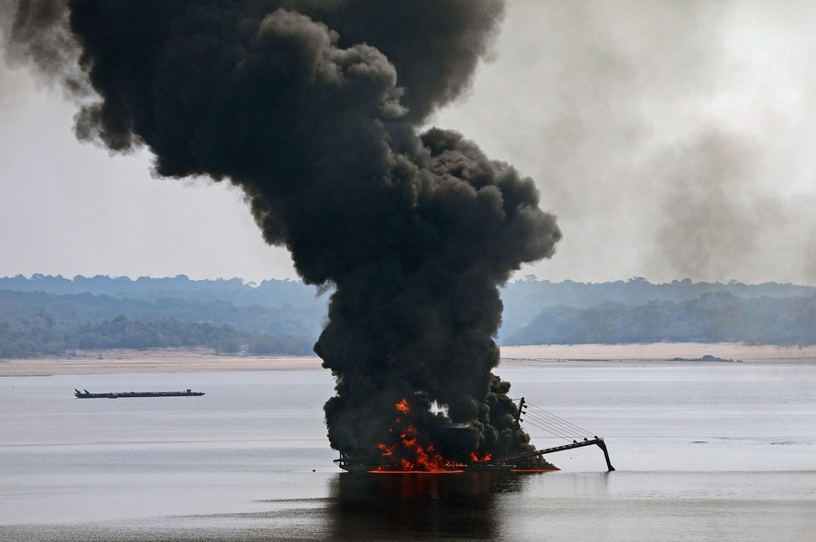 A floating piece of mining equipment burns in a river, sending up a column of black smoke.
