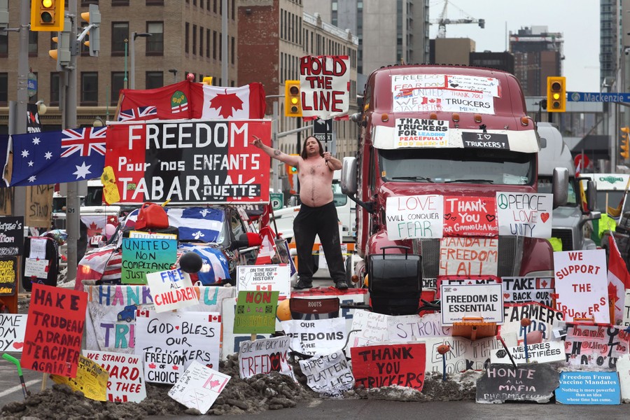 A shirtless person stands next to a truck, surrounded by dozens of protest signs, on a city street.