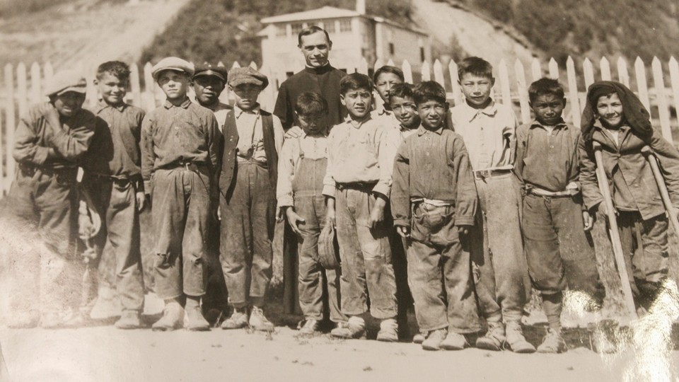 About a dozen Indigenous boys stand in front of a priest, all facing the camera.